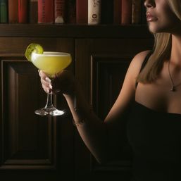 Woman in a black dress holding a bright yellow cocktail in a coupe glass with a kiwi slice, moody wood‑paneled bookshelf backdrop, dim bar lighting.