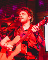 Red-lit live music scene: curly-haired singer strumming an acoustic guitar and singing into a mic on an intimate indoor bar/club stage.