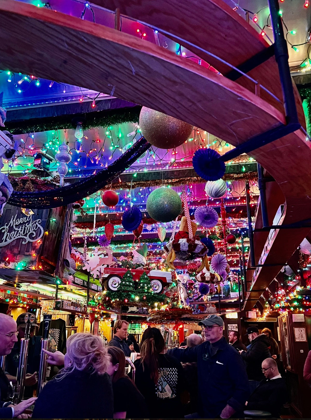 Holiday-themed crowded pub interior with multicolored string lights, giant hanging ornaments and paper decorations, a suspended red toy car above a busy bar and patrons chatting.