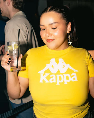 Person in a bright yellow Kappa t-shirt smiling with eyes closed at a bar, holding a tall lime cocktail with a black straw, wearing dangling cross earring and black nail polish — nightlife party vibe.