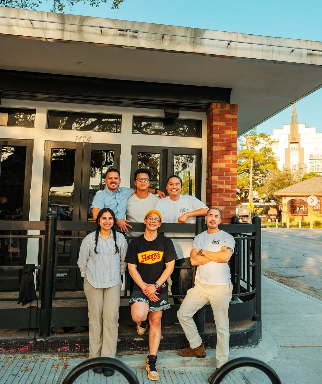 Six smiling people pose together outside a brick-front neighborhood cafe on a sunny city street, leaning on a black railing with trees and blue sky behind them.
