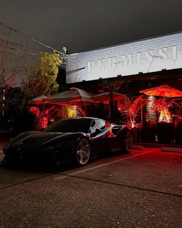 Black luxury sports car parked at night outside a red-lit urban restaurant patio with palm plants, umbrellas and illuminated brick signage.