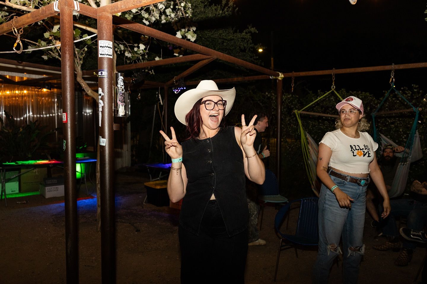 Smiling woman in a white cowboy hat and glasses flashing two peace signs at a nighttime outdoor patio party, friends nearby in jeans and caps with hammocks and string lights in the background.