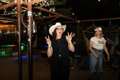 Smiling woman in a white cowboy hat and glasses flashing two peace signs at a nighttime outdoor patio party, friends nearby in jeans and caps with hammocks and string lights in the background.
