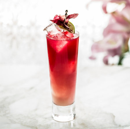 Refreshing layered red-to-pink iced cocktail in a tall highball glass, garnished with a lime slice and hibiscus flower on a marble surface