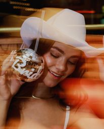 Smiling woman in a white cowboy hat holding a mirrored disco-ball drink cup labeled "Bride" with a straw, warm club lights and motion blur for a bachelorette party vibe