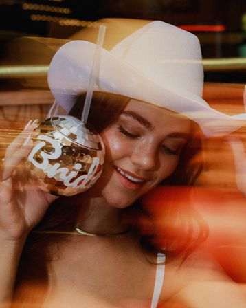 Smiling woman in a white cowboy hat holding a mirrored disco-ball drink cup labeled "Bride" with a straw, warm club lights and motion blur for a bachelorette party vibe