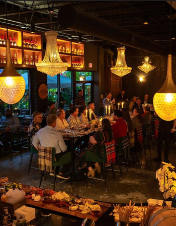 Cozy upscale indoor dinner scene with a long communal table of guests, glowing crystal chandeliers and candlelight, and a charcuterie board in the foreground under warm ambient lighting.