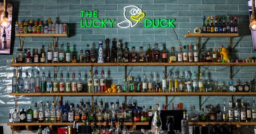 Green neon duck sign glowing above a trendy bar with teal subway-tile wall, wooden shelves packed with liquor bottles, glassware and playful rubber ducks.