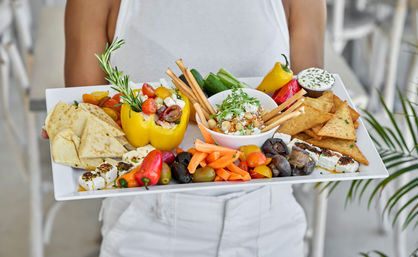Vibrant Mediterranean mezze platter on a white tray: hummus with breadsticks, pita triangles, feta cubes, olives, stuffed yellow pepper, carrot sticks, mushrooms and pita chips, held by a person in white.
