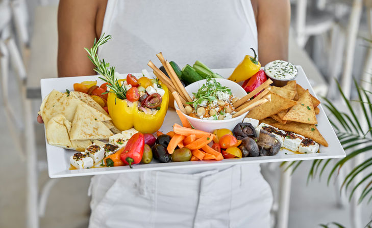 Vibrant Mediterranean mezze platter on a white tray: hummus with breadsticks, pita triangles, feta cubes, olives, stuffed yellow pepper, carrot sticks, mushrooms and pita chips, held by a person in white.