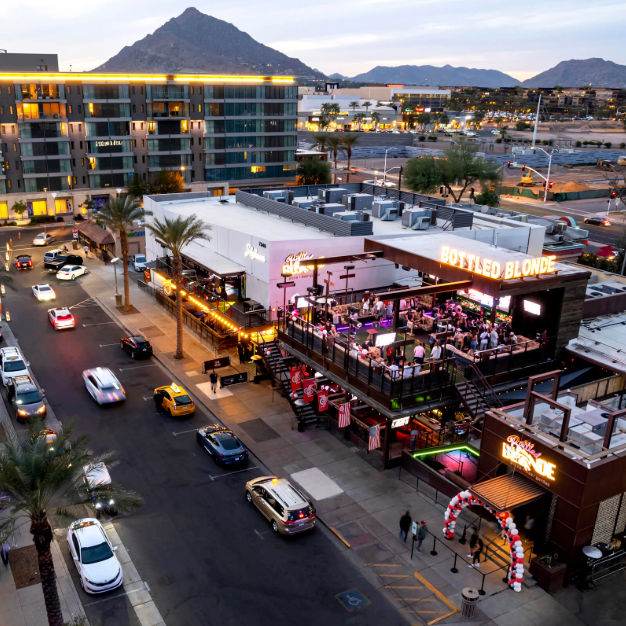 Aerial view of a buzzing rooftop bar and outdoor patio packed with people on a downtown street at dusk, palm trees and cars below, modern apartments and rugged mountains silhouetted in the background.
