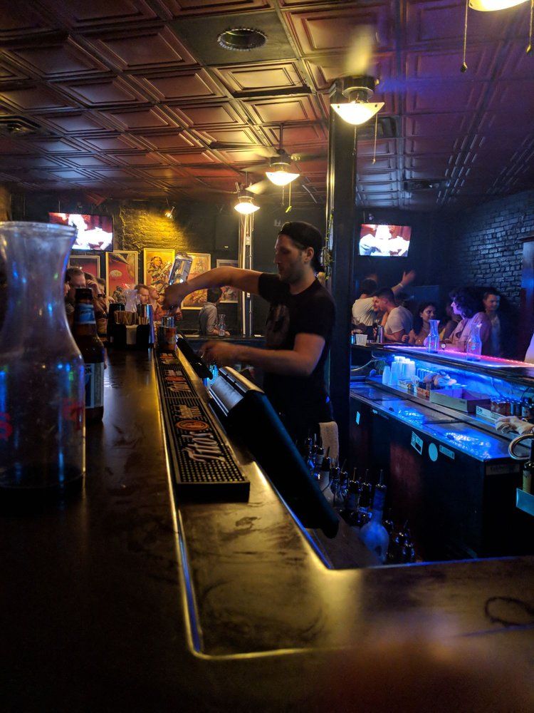 Bartender pouring drinks at a dimly lit indoor bar with tin ceiling, neon blue backlit counter and patrons socializing — lively nightlife scene.