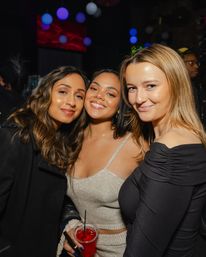 Three smiling women posing at a lively nightclub party, one holding a red cocktail under colorful hanging lights — fun urban nightlife vibe