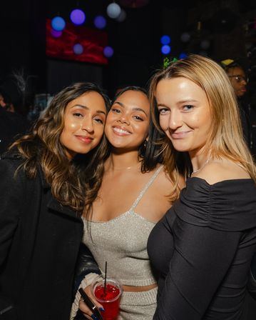 Three smiling women posing at a lively nightclub party, one holding a red cocktail under colorful hanging lights — fun urban nightlife vibe