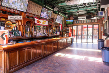 Sunlit sports bar interior with a long polished wooden bar, brick walls, exposed green ductwork, multiple flat-screen TVs, beer taps, tiled floor and open glass doors to an outdoor patio.
