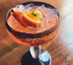 Vibrant pink grapefruit cocktail in a coupe glass with crushed ice, orange peel and microgreen garnish, chili-salt rim, resting on a wooden bar