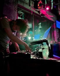 DJ mixing on a controller with a laptop and drink on a table in a neon-lit urban nightclub, pink and green lights and smoky vibe