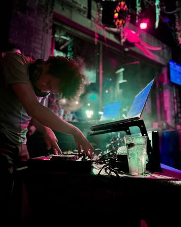 DJ mixing on a controller with a laptop and drink on a table in a neon-lit urban nightclub, pink and green lights and smoky vibe