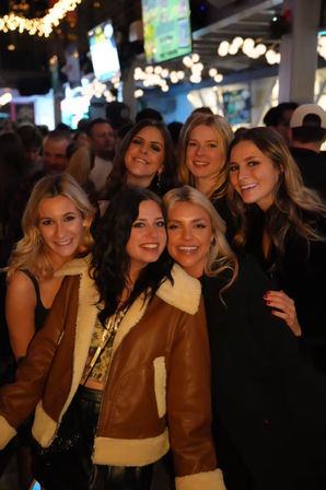 Six friends smiling for a group photo during a lively night out at a downtown bar with string lights and TVs in the background