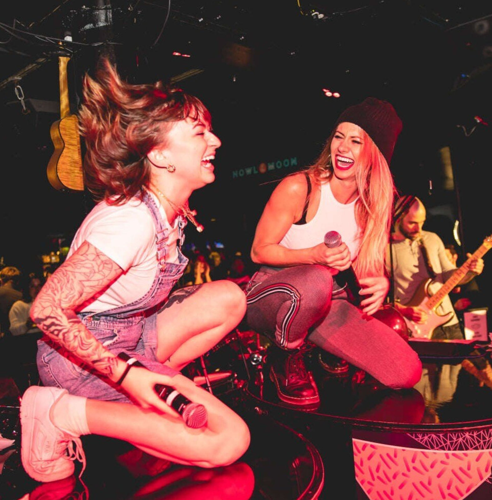 Two singers crouch on a bar-stage laughing and holding microphones under pink stage lights, a guitarist plays behind them at a packed live-music venue.