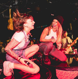 Two singers crouch on a bar-stage laughing and holding microphones under pink stage lights, a guitarist plays behind them at a packed live-music venue.