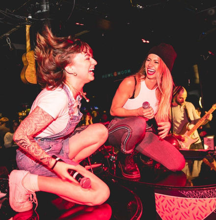 Two singers crouch on a bar-stage laughing and holding microphones under pink stage lights, a guitarist plays behind them at a packed live-music venue.