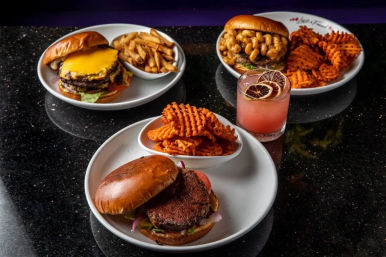 Three gourmet burgers on white plates with bowls of fries and sweet potato waffle fries — a cheddar-topped cheeseburger, a mac-and-cheese–topped burger, and a crispy patty — plus a pink citrus cocktail on a black granite table.