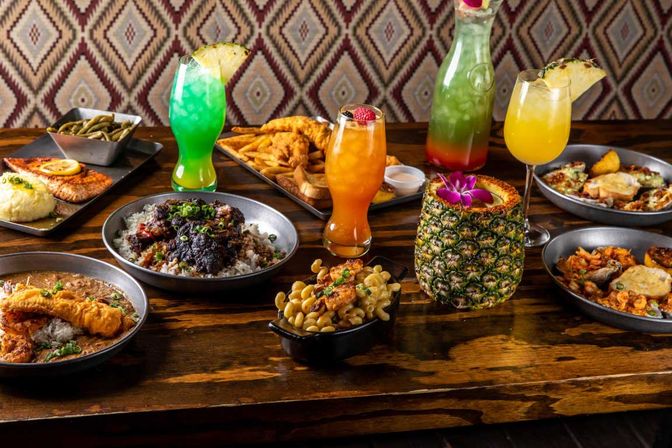 Island-inspired table spread: hollowed pineapple cocktail with orchid, bright green and orange tropical drinks, pitcher, fried fish over rice, mac and cheese, fries and seafood plates on a rustic wooden table