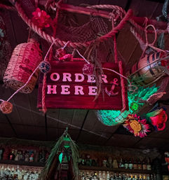 Tiki bar ceiling with a hanging wooden "ORDER HERE" sign surrounded by ropes, fishing nets, buoys, a faux pineapple and colorful tropical flowers above a shelf of liquor bottles — nautical bar decor.