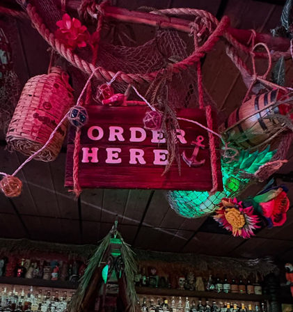 Tiki bar ceiling with a hanging wooden "ORDER HERE" sign surrounded by ropes, fishing nets, buoys, a faux pineapple and colorful tropical flowers above a shelf of liquor bottles — nautical bar decor.