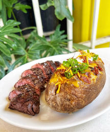 Sliced grilled steak with pepper crust served alongside a loaded baked potato topped with melted cheddar, bacon, sour cream and green onions on a white plate, leafy plants and a yellow wall in the background.