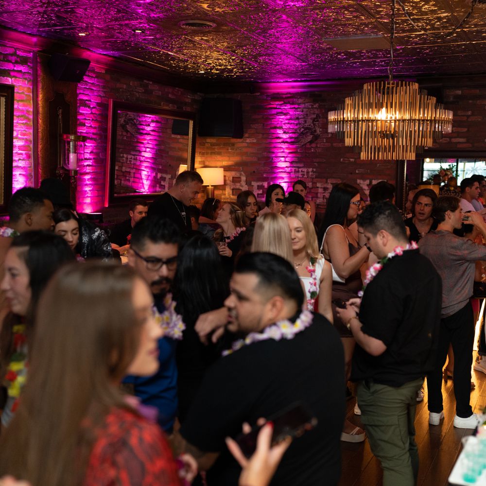 Lively urban lounge with exposed brick, pink uplighting and a gold chandelier, crowded with people mingling and wearing floral leis at a nighttime party.