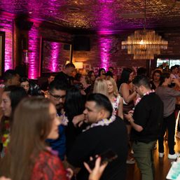 Lively urban lounge with exposed brick, pink uplighting and a gold chandelier, crowded with people mingling and wearing floral leis at a nighttime party.