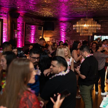 Lively urban lounge with exposed brick, pink uplighting and a gold chandelier, crowded with people mingling and wearing floral leis at a nighttime party.