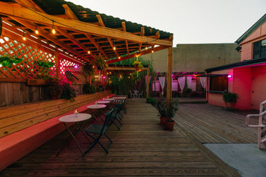 Cozy outdoor wooden-deck patio with a pergola and hanging string lights, bistro tables and folding chairs along a bench, potted plants and curtained lounge cabanas glowing with pink evening light.