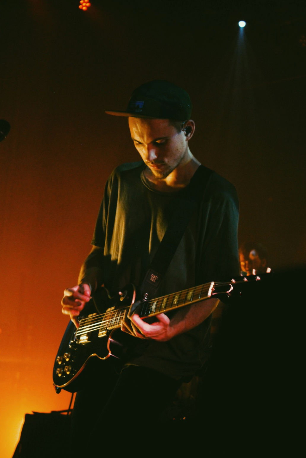 Guitarist playing a black electric guitar at a live indoor concert, bathed in warm orange stage light and wearing a cap while focused on the performance.