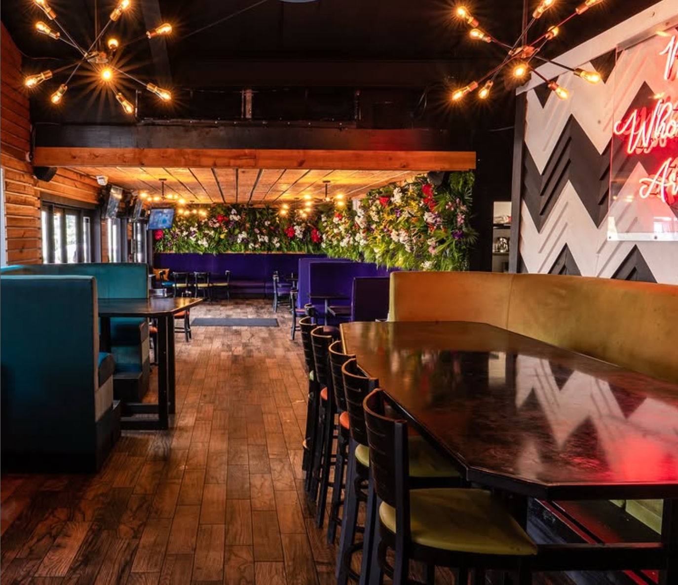 Modern restaurant interior with wooden floor, teal booths and long high-top table with bar stools, purple banquette seating and a lush living floral wall under warm exposed-bulb pendant lighting and chevron accent wall.