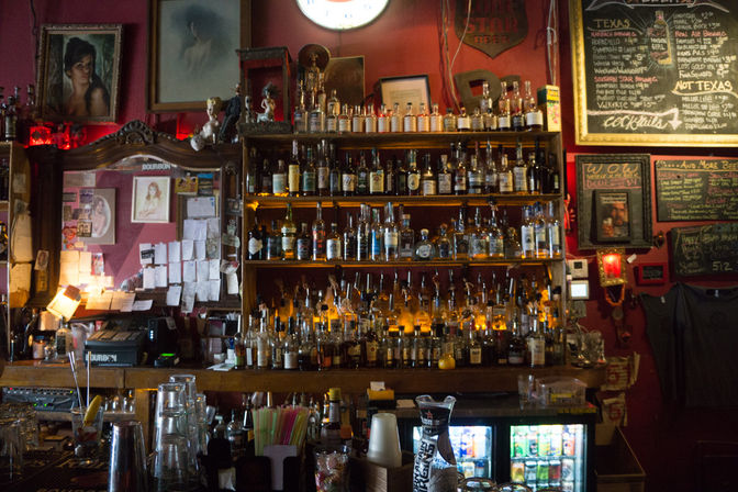 Cozy dimly lit bar interior with backlit wooden shelves of liquor bottles, chalkboard menu, vintage mirror and a cluttered counter with glasses, straws and mixers.