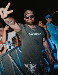 Smiling tattooed festival-goer in a cap and sunglasses flashes a peace sign from the front-row barrier at an outdoor night music festival, crowd and wristbands visible