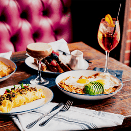 Vibrant brunch spread on a rustic wooden table in a cozy cafe — herb-topped omelette, sunny-side eggs with sliced avocado, berry-topped toast, espresso cocktail and a spritz.