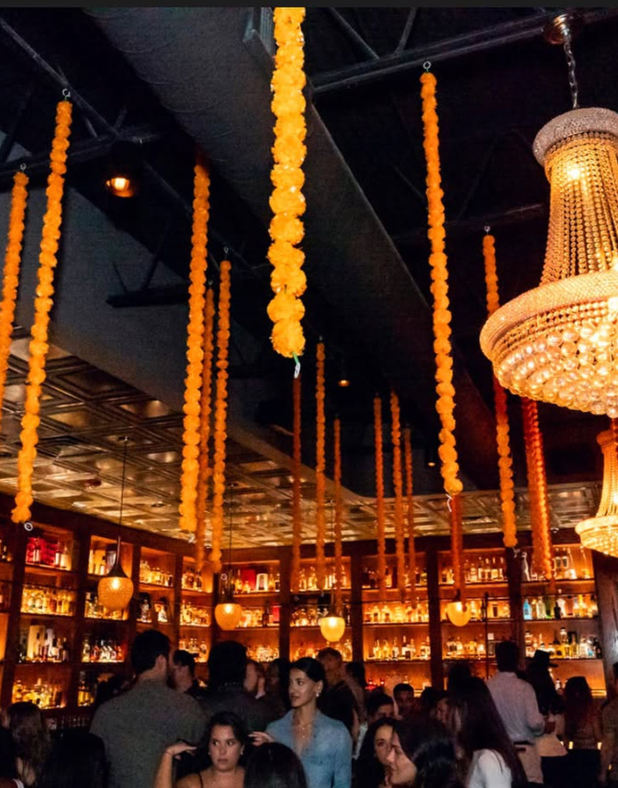 Festive upscale bar interior with warm backlit liquor shelves, a crystal chandelier, and long orange marigold-style garlands hanging from the ceiling above a lively evening crowd.