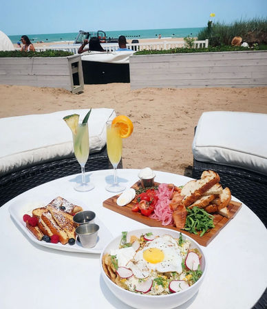 Beachfront brunch spread on a white table: two tropical mimosas, French toast with berries, a wooden charcuterie board with smoked salmon and toasted bread, a bowl topped with a sunny-side egg, lounge chairs and turquoise ocean in the background