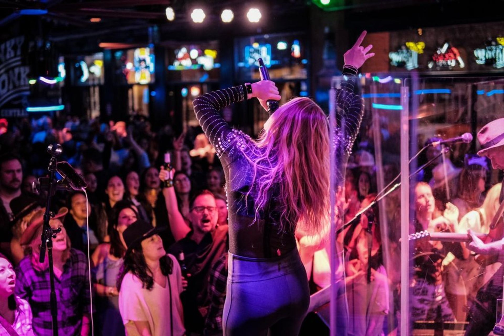 Lead singer with long hair on a neon-lit stage, microphone raised and one arm in the air as a packed, cheering crowd enjoys live music while a guitarist plays stage right
