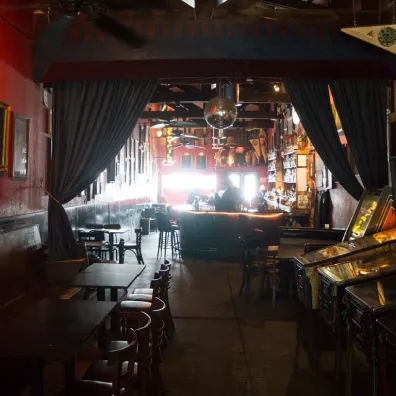 Dimly lit, narrow bar interior with wooden tables and stools, disco ball overhead, retro pinball machines and a glowing backlit bar counter.