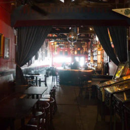 Dimly lit, narrow bar interior with wooden tables and stools, disco ball overhead, retro pinball machines and a glowing backlit bar counter.
