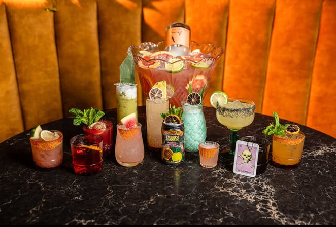 Assorted colorful craft cocktails and a large fruit-filled punch bowl arranged on a black marble table in a warm orange velvet lounge, each drink garnished with citrus, mint, dried fruit, and spiced-rimmed glasses.