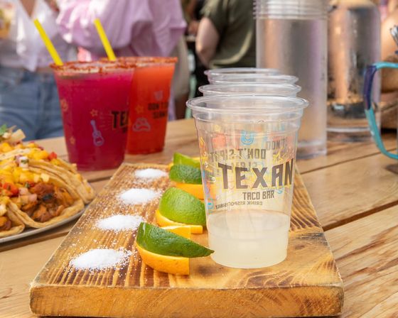 Wooden tasting board with lime and orange wedges and salt piles beside plastic sample cups of margarita and bright cocktails, with tacos on a plate on a sunny outdoor patio
