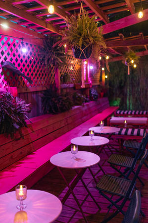 Cozy neon-pink alfresco patio at night under a wooden pergola with hanging fern planters, string lights, candlelit round bistro tables and folding chairs