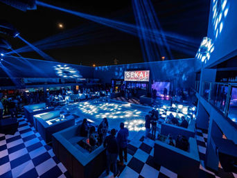Rooftop nightclub and outdoor lounge at night with checkerboard floor, blue LED beams, illuminated central dance floor and VIP booths with guests